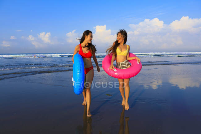 Two pretty girls with floaties on the way to the waves of the ocean. One girl is turning around smiling. — Stock Photo