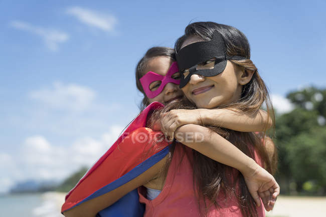 Giovane madre asiatica con carina figlia in costumi da supereroe in posa contro il cielo blu — Foto stock