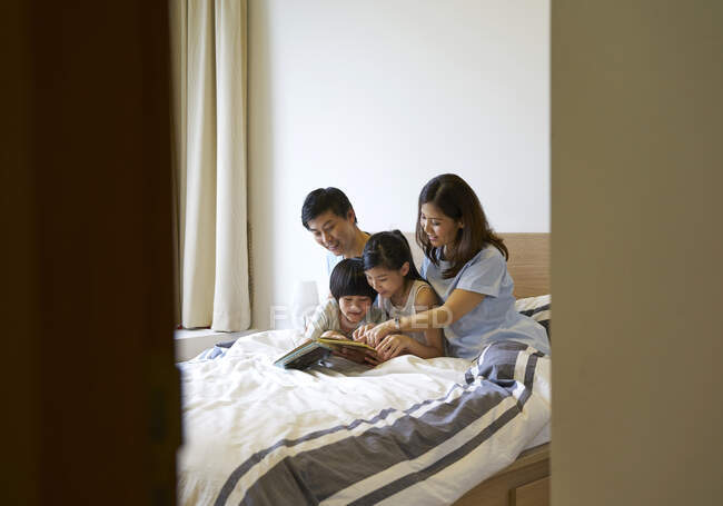 Felice giovane famiglia asiatica insieme lettura libro in camera da letto — Foto stock