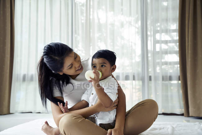 Asiático madre relojes como su hijo se alimenta de leche botella - foto de stock