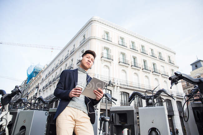 Hombre chino joven casual con una tableta. auriculares y una taza de café en Madrid, España - foto de stock