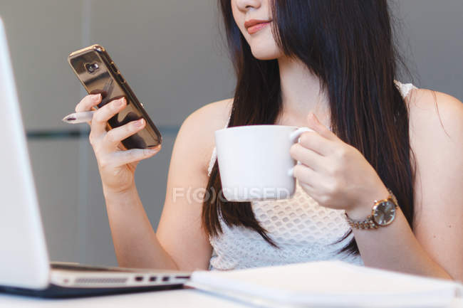 Mujer joven revisando su teléfono con café en la mano en la oficina moderna - foto de stock