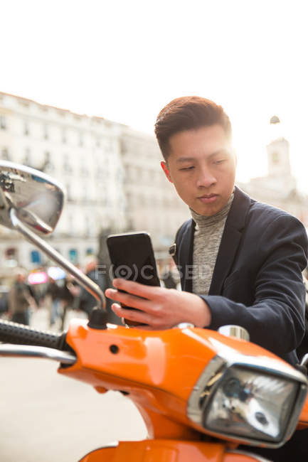 Casual jovem chinês com um telefone inteligente. sentado em uma moto na Puerta del Sol, Madrid, Espanha — Fotografia de Stock
