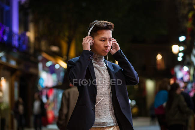 Casual jovem chinês com fones de ouvido pendurados nas ruas de Madrid à noite, Espanha — Fotografia de Stock