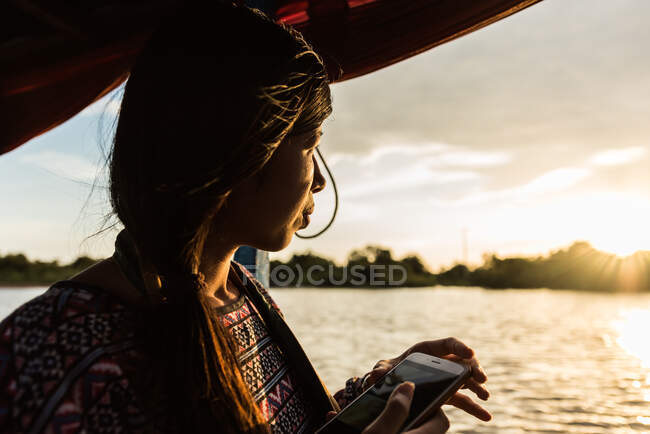Junge Frau sitzt in einem Boot und betrachtet den Sonnenuntergang, während sie ein Telefon in der Hand hält, mit einem Lächeln im Gesicht — Stockfoto