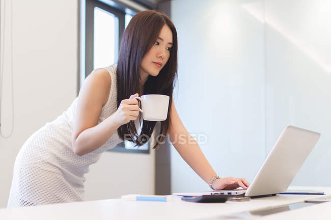 Jeune femme vérifiant son ordinateur portable avec tasse de café dans le bureau moderne — Photo de stock