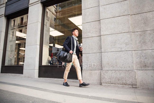 Hombre chino joven casual caminando por la calle sosteniendo una taza de café, España - foto de stock