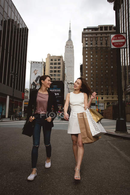Two beautiful elegant women passing street and holding shopping bags with empire state behind. — Stock Photo