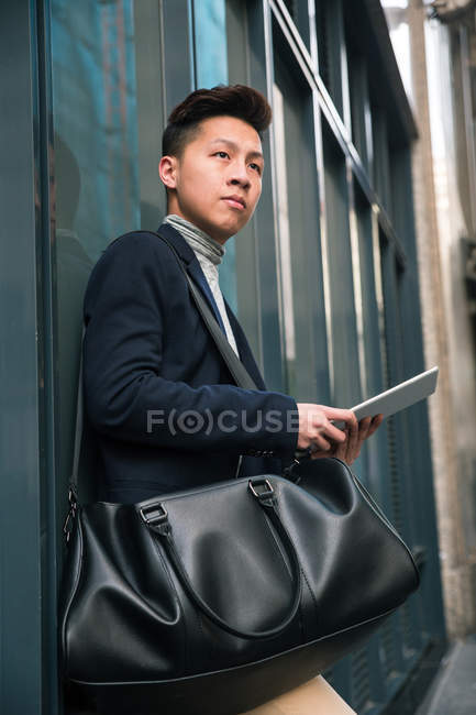Lässiger junger chinesischer Mann schaut weg und hält eine Tablette in Madrid, Spanien — Stockfoto