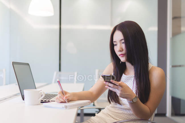 Jeune femme prenant des notes avec le téléphone dans la main dans le bureau moderne — Photo de stock