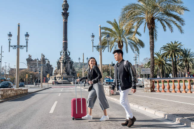 Jeune couple touristique traversant la route au monument de Colomb avec une valise — Photo de stock