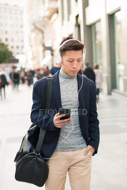 Casual young chinese man using phone and headphones in the street — Stock Photo
