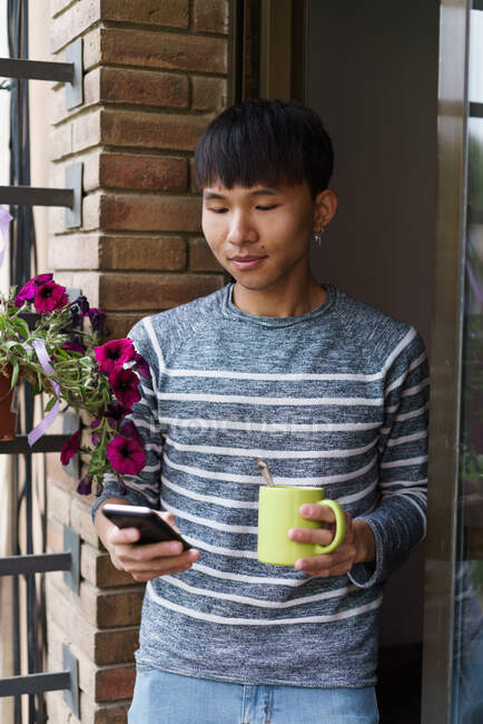 Молодой азиат улыбающийся мужчина держит кружку с кофе и просматривает смартфон, стоя на балконе. — стоковое фото