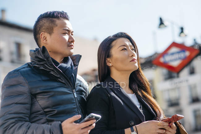 Turistas chinos asiáticos de luna de miel caminando por la plaza de la ópera y el teatro mirando sus teléfonos móviles en Madrid - foto de stock