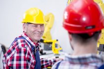 Homem na fábrica usando chapéu duro sorrindo para colega — Fotografia de Stock