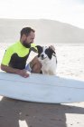 Hombre con perro y tabla de surf sentado en la playa de arena - foto de stock
