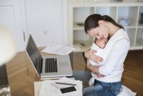 Seitenansicht der freiberuflichen Mutter, die ihr Kind im Tragetuch im Home Office umarmt — Stockfoto