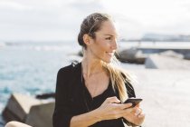 Lächelnde junge Frau mit Handy am Meer, die sich umschaut — Stockfoto