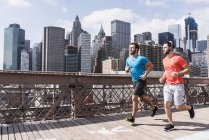Estados Unidos, Nueva York, puente de Brooklyn, dos jóvenes trotando, vista del paisaje urbano en segundo plano - foto de stock