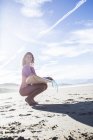 Young woman with surfboard sitting at sandy beach smiling looking aside, sunny seascape on background — Stock Photo