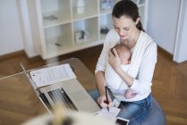 Freiberufliche Mutter mit Säugling im Tragetuch macht sich Notizen im Home Office — Stockfoto