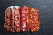 Top view of arranged slices of ham and salami on black schist background — Stock Photo