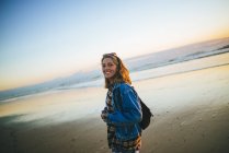 Young woman walking on the beach at sunset, looking at camera — Stock Photo