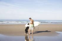 Homem com o cão segurando prancha na praia arenosa — Fotografia de Stock