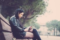 Giovane donna dai capelli scuri seduta sulla panchina e che ascolta musica con auricolari e smartphone — Foto stock