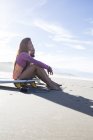 Young woman with surfboard sitting at sandy beach looking aside — Stock Photo