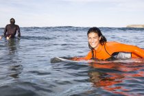 Mulher deitada na prancha de surf no mar sorrindo para a câmera, visão traseira do homem na prancha de surf à espera de onda no fundo — Fotografia de Stock