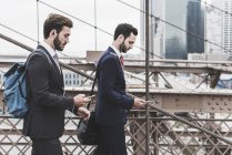 США, Нью-Йорк, Бруклінський міст, двоє бізнесменів з смартфонів і навушниками ходити в місті — стокове фото