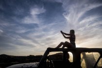 Silhouette di donna seduta sul tetto dell'auto al tramonto e scattare foto — Foto stock