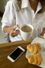 Woman with coffee, croissants and cell phone at breakfast table — Stock Photo