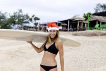 Lächelnde Frau mit Surfbrett am Strand mit Weihnachtsmütze — Stockfoto