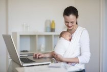 Mère pigiste avec bébé en fronde travaillant au bureau à domicile — Photo de stock