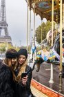 Paris, France, deux meilleurs amis utilisant leur smartphone avec un carrousel et la Tour Eiffel en arrière-plan
. — Photo de stock