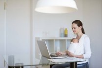 Visão lateral da mãe freelancer com bebê em sling trabalhando no escritório em casa — Fotografia de Stock