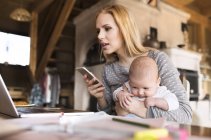 Mother with baby using laptop and cell phone at home — Stock Photo