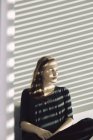 Портрет жінка, що сидить у смугастих sunshine позаду жалюзі, дивлячись за межами — стокове фото