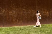 Portrait de jeune femme marchant avec téléphone portable et sac à bandoulière près du mur brun — Photo de stock