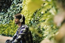 Young man sitting in front of green plant wall, wearing head phones — Stock Photo