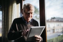 Uomo anziano utilizzando tablet a casa rilassato, vicino alla finestra
. — Foto stock