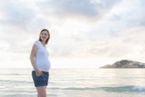 Mujer embarazada feliz de pie en la playa con el cielo nublado en el fondo - foto de stock