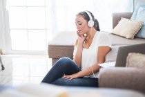Mujer joven sentada en el suelo de la sala de estar escuchando música con auriculares - foto de stock