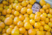 Little smiling boy lying under pile of oranges — Stock Photo