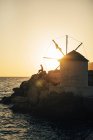 Grecia, Amorgos, Aegialis, silueta del hombre sentado cerca del molino de viento al atardecer - foto de stock