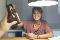 Усміхаючись дзвінкою пляшку пива жінка сидить за столом під ліхтарем — стокове фото