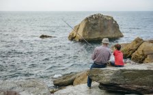 Abuelo y nieto pescando juntos en el mar sentado en la roca - foto de stock