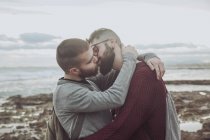 Joven pareja gay abrazándose y besándose delante del mar - foto de stock
